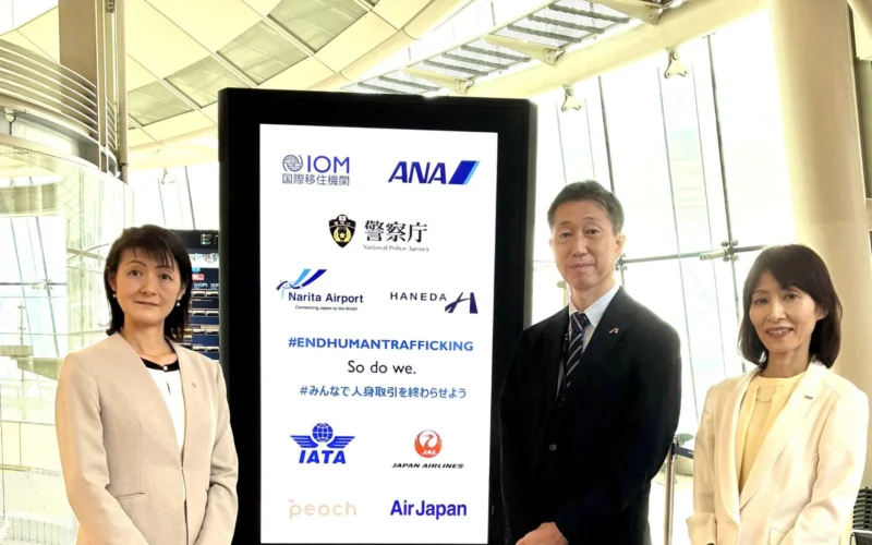 Three professionals in light colored suits posing beside a tall digital display board filled with airline logos and an anti trafficking message at an airport terminal