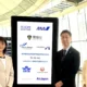Three professionals in light colored suits posing beside a tall digital display board filled with airline logos and an anti trafficking message at an airport terminal