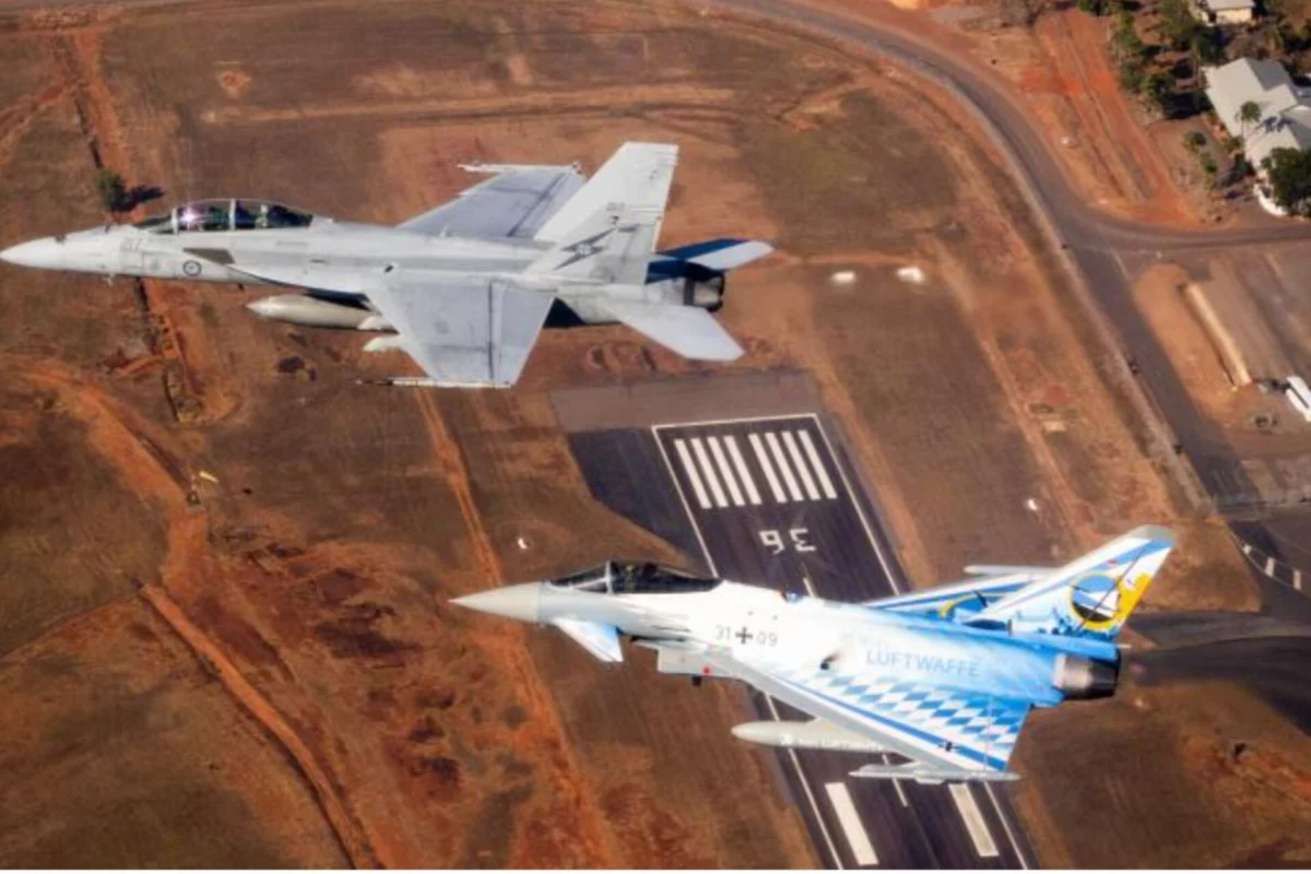 Two military fighter jets flying in formation over a paved airstrip at an arid airbase