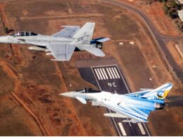 Two military fighter jets flying in formation over a paved airstrip at an arid airbase