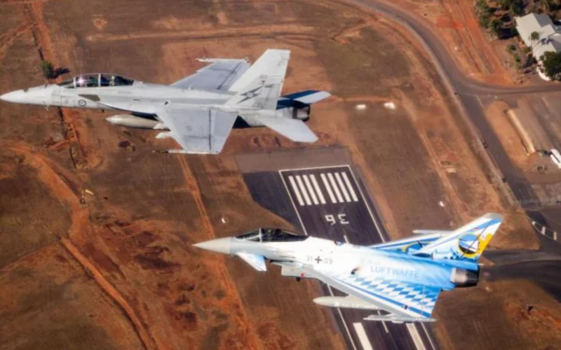 Two military fighter jets flying in formation over a paved airstrip at an arid airbase