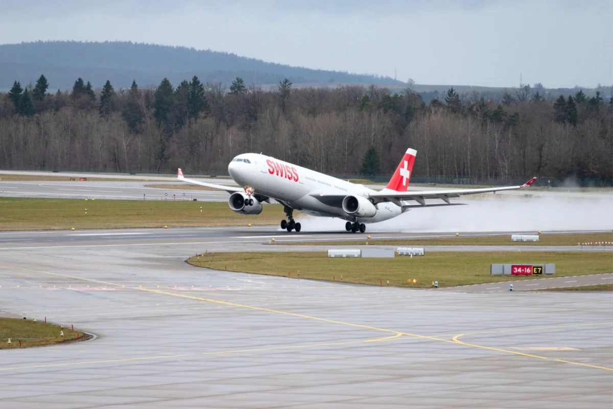 Swiss aircraft lifting off a rainy runway spray trailing from the wheels as it begins takeoff with forested hills in the background