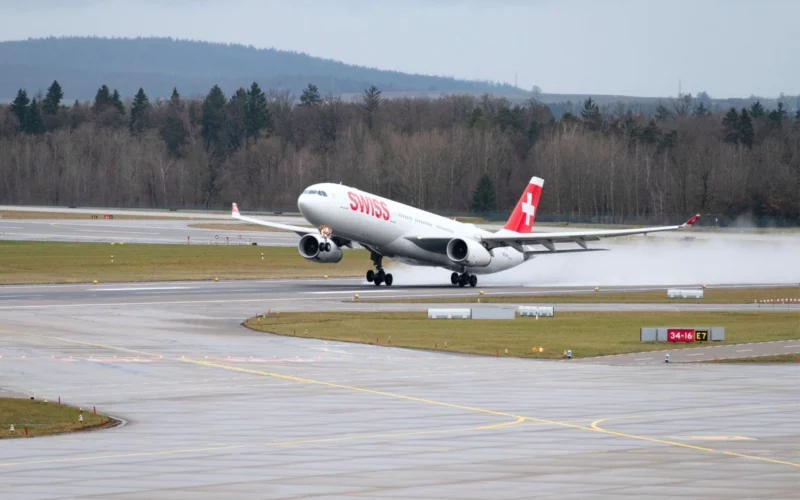 Swiss aircraft lifting off a rainy runway spray trailing from the wheels as it begins takeoff with forested hills in the background