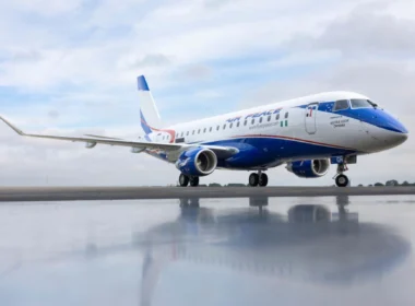 Airplane on a runway with blue nose and tail, reflection visible on the wet tarmac under a cloudy sky.
