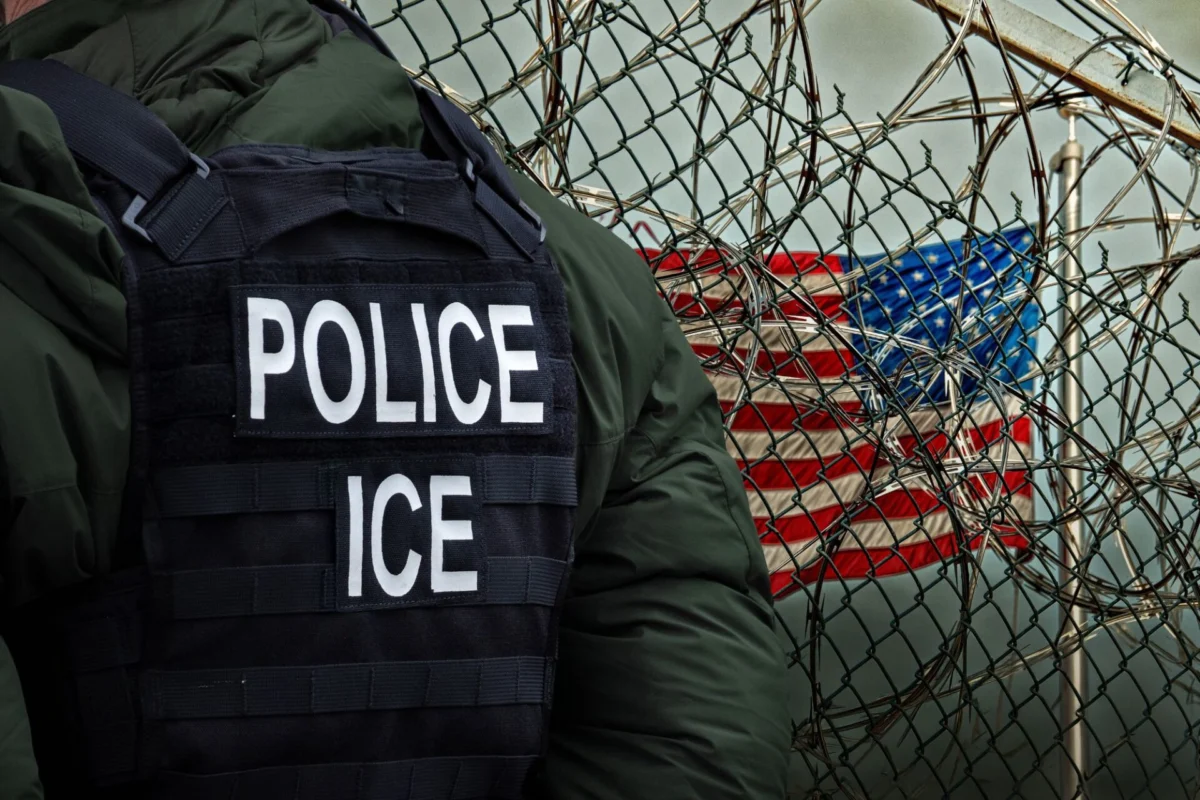 Officer wearing a tactical vest labeled POLICE and ICE beside a barbed wire fence with the US flag in the background conveying law enforcement near a barrier