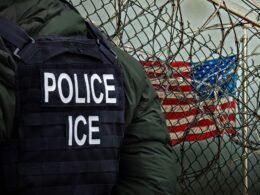 Officer wearing a tactical vest labeled POLICE and ICE beside a barbed wire fence with the US flag in the background conveying law enforcement near a barrier