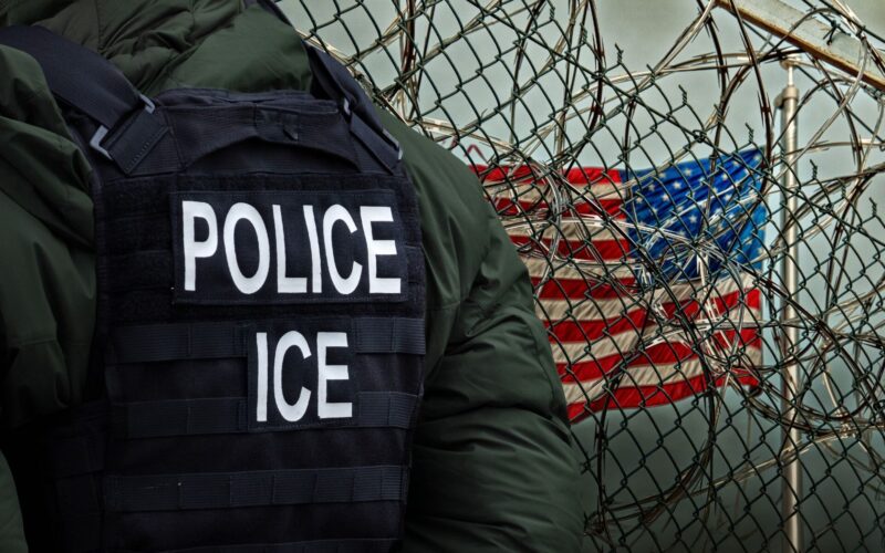 Nonprofit watchdog releases TSA ICE data agreement Officer wearing a tactical vest labeled POLICE and ICE beside a barbed wire fence with the US flag in the background conveying law enforcement near a barrier