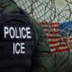Officer wearing a tactical vest labeled POLICE and ICE beside a barbed wire fence with the US flag in the background conveying law enforcement near a barrier