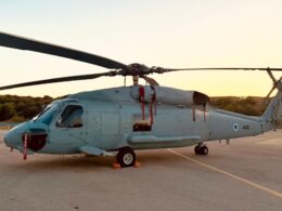 Side profile of a gray military helicopter parked on a tarmac at sunset rotor blades extended to the sides