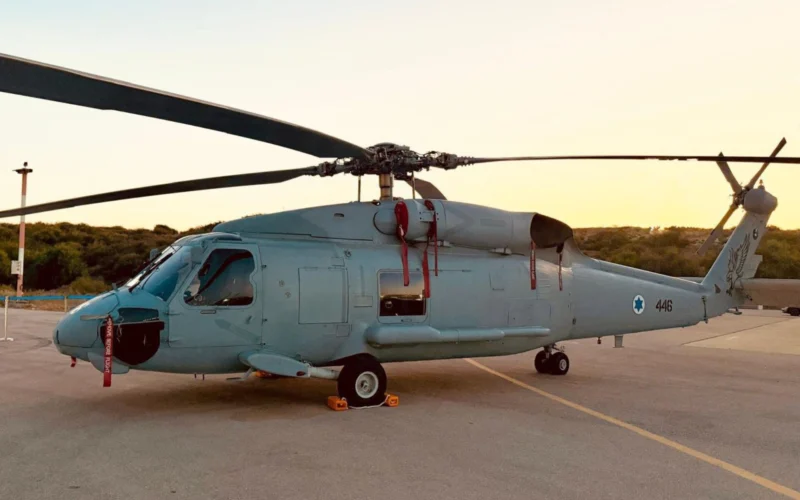 Side profile of a gray military helicopter parked on a tarmac at sunset rotor blades extended to the sides