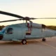 Side profile of a gray military helicopter parked on a tarmac at sunset rotor blades extended to the sides
