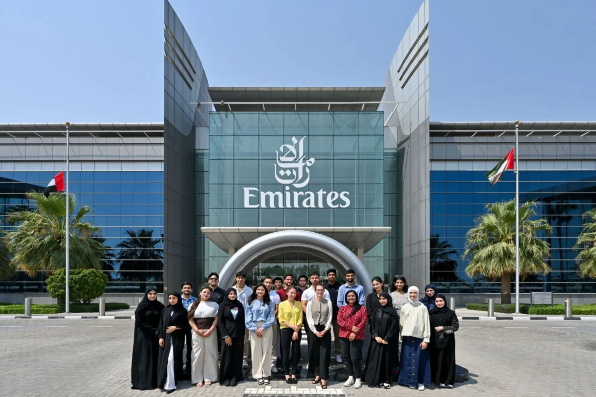 Group of diverse people posing for a photo in front of a modern Emirates building with glass facade and flags