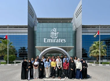 Group of diverse people posing for a photo in front of a modern Emirates building with glass facade and flags.