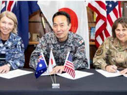 Three military officers in uniform signing documents at a table with small flag flags Australia UK Japan USA in front and national flags displayed behind them