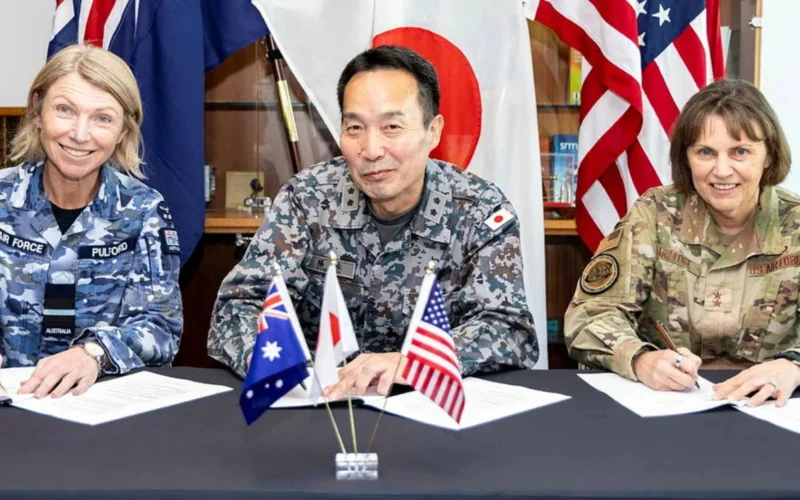 Three military officers in uniform signing documents at a table with small flag flags Australia UK Japan USA in front and national flags displayed behind them