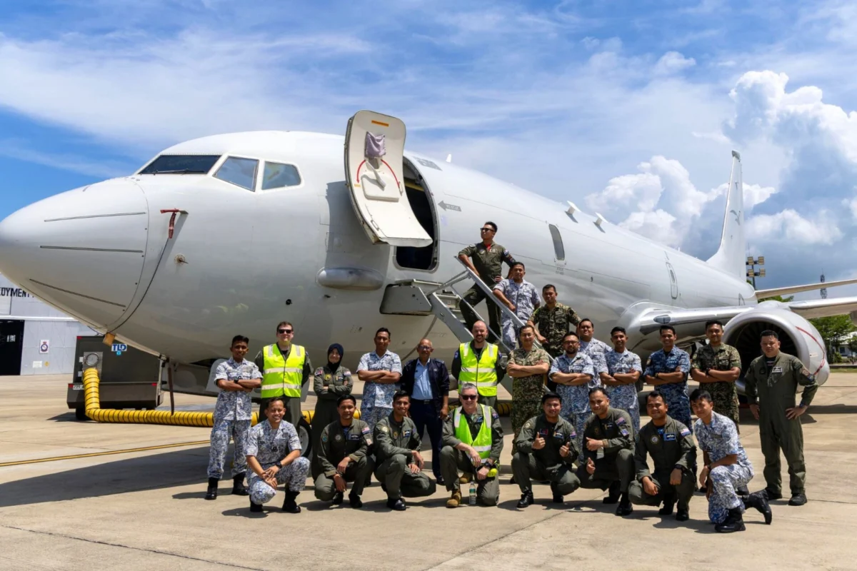 Group of diverse military personnel posing in front of a large white passenger jet with its door open on the tarmac at a sunny day airport