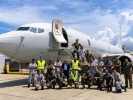 Group of diverse military personnel posing in front of a large white passenger jet with its door open on the tarmac at a sunny day airport