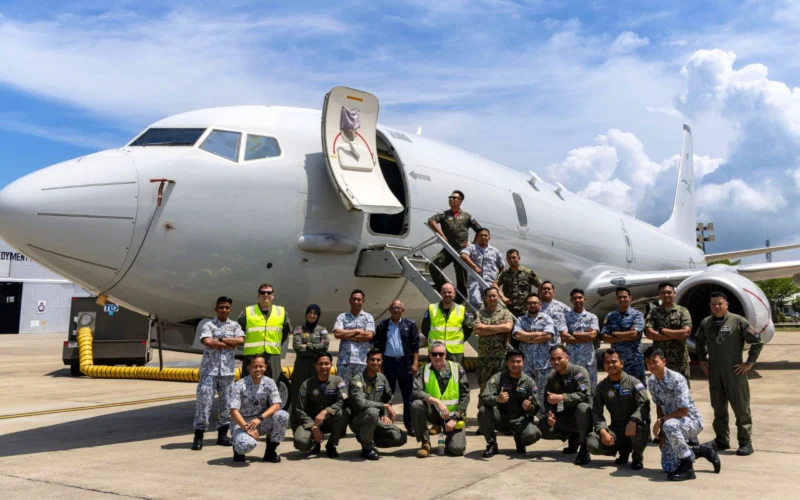 Group of diverse military personnel posing in front of a large white passenger jet with its door open on the tarmac at a sunny day airport