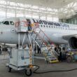 Inside a large aircraft hangar a Lufthansa CityLine jet is being serviced with orange maintenance stairs and ground crews nearby in a bright open space