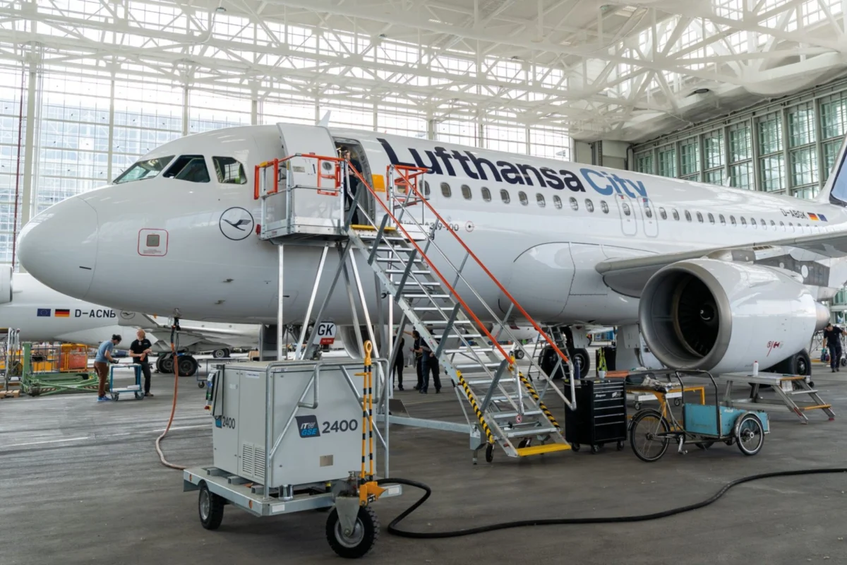 Inside a large aircraft hangar a Lufthansa CityLine jet is being serviced with orange maintenance stairs and ground crews nearby in a bright open space