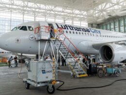 Inside a large aircraft hangar a Lufthansa CityLine jet is being serviced with orange maintenance stairs and ground crews nearby in a bright open space