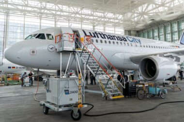 Inside a large aircraft hangar a Lufthansa CityLine jet is being serviced with orange maintenance stairs and ground crews nearby in a bright open space