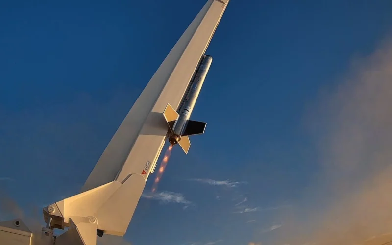 Rocket mounted to a white aircraft tail during a test launch flames trailing from its engine against a blue sky