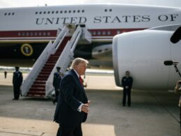 Man in a dark suit and red tie walks on the tarmac beside a large white aircraft labeled United States as media crew film nearby