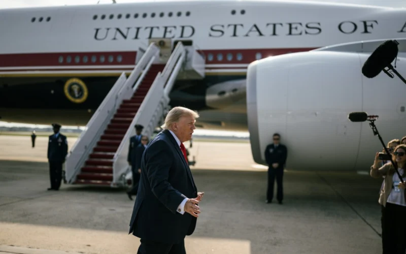 Man in a dark suit and red tie walks on the tarmac beside a large white aircraft labeled United States as media crew film nearby