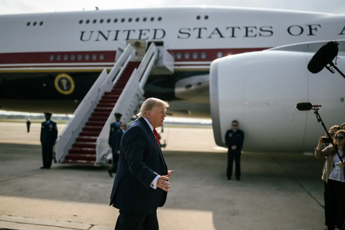 af1 Man in a dark suit and red tie walks on the tarmac beside a large white aircraft labeled United States as media crew film nearby