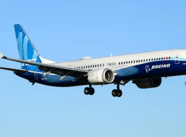 Blue and white Boeing 737 MAX in flight with landing gear extended against a clear blue sky