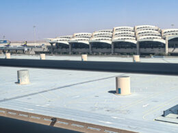 View of a large airport terminal with curved multi level structures in the distance and a spacious empty rooftop parking lot in the foreground under a clear blue sky