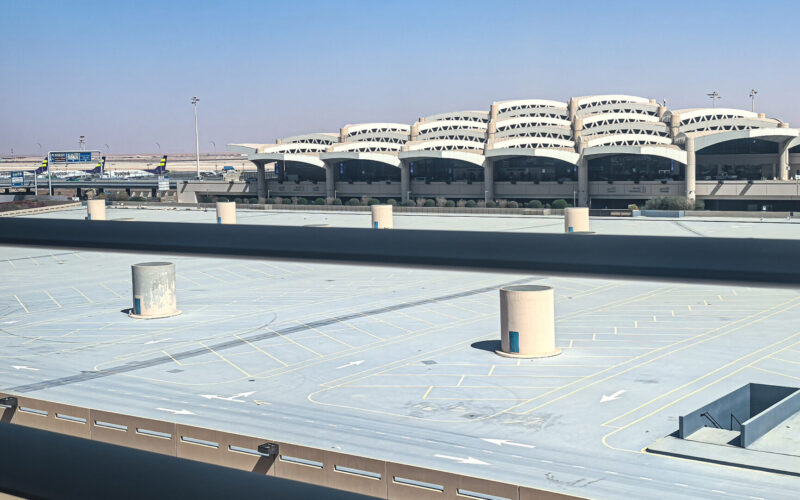 airport king khalid international View of a large airport terminal with curved multi level structures in the distance and a spacious empty rooftop parking lot in the foreground under a clear blue sky
