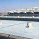 View of a large airport terminal with curved multi level structures in the distance and a spacious empty rooftop parking lot in the foreground under a clear blue sky