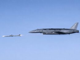 Gray stealth bomber in flight releasing a missile with a visible exhaust flame against a clear blue sky