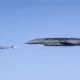 Gray stealth bomber in flight releasing a missile with a visible exhaust flame against a clear blue sky