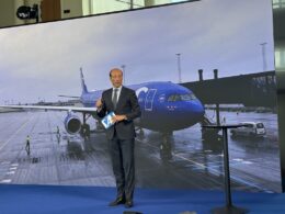 Business presenter in a suit stands on a blue stage holding a blue SA card with a large screen showing a blue airplane at an airport behind him