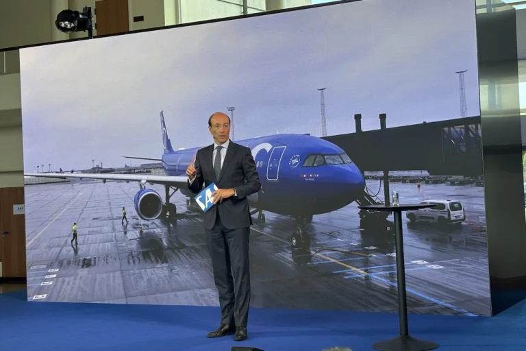 Business presenter in a suit stands on a blue stage holding a blue SA card with a large screen showing a blue airplane at an airport behind him