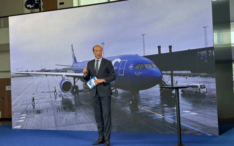 Business presenter in a suit stands on a blue stage holding a blue SA card with a large screen showing a blue airplane at an airport behind him