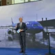 Business presenter in a suit stands on a blue stage holding a blue SA card with a large screen showing a blue airplane at an airport behind him