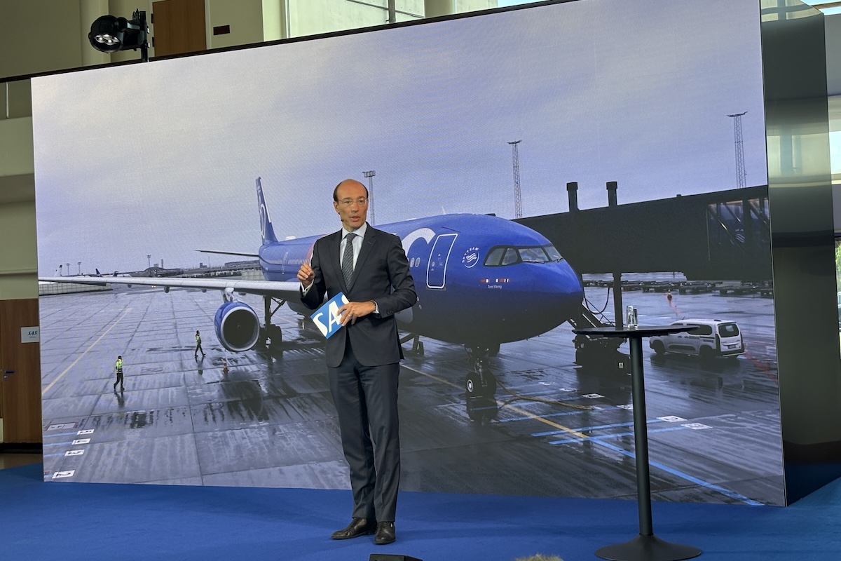 Business presenter in a suit stands on a blue stage holding a blue SA card with a large screen showing a blue airplane at an airport behind him