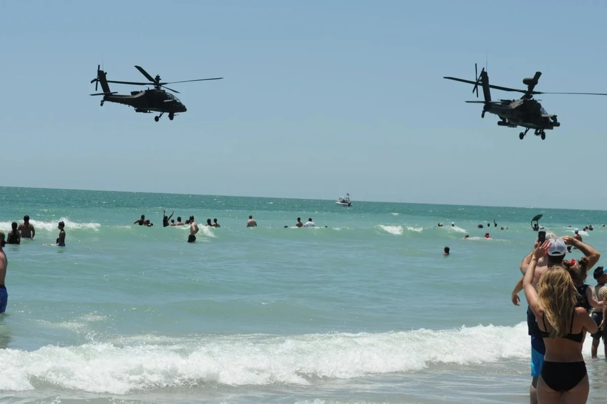 apache flyby 3x2 Two military helicopters fly low over a crowded sunny beach as people swim and play in the waves on a bright day near the shore