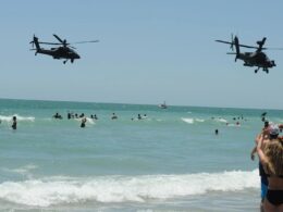 Two military helicopters fly low over a crowded sunny beach as people swim and play in the waves on a bright day near the shore