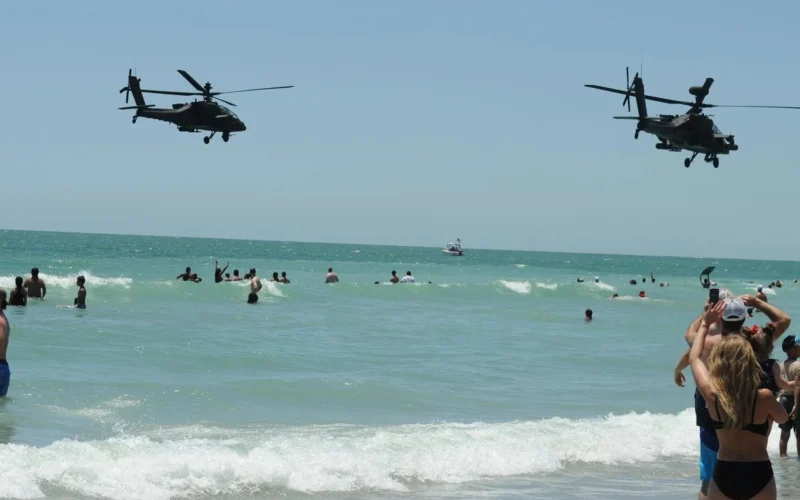Two military helicopters fly low over a crowded sunny beach as people swim and play in the waves on a bright day near the shore