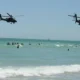 Two military helicopters fly low over a crowded sunny beach as people swim and play in the waves on a bright day near the shore