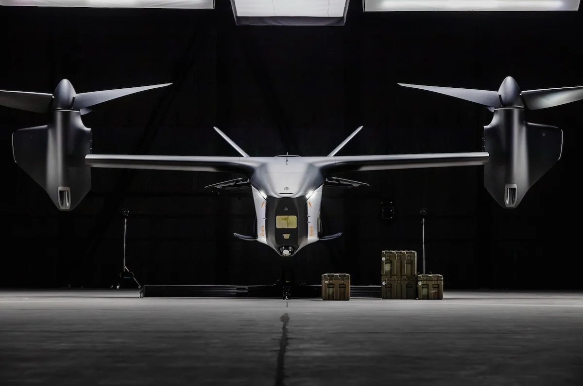 archer anduril Front on view of a sleek silver eVTOL aircraft in a dark hangar with cargo cases on the floor nearby