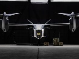 Front on view of a sleek silver eVTOL aircraft in a dark hangar with cargo cases on the floor nearby