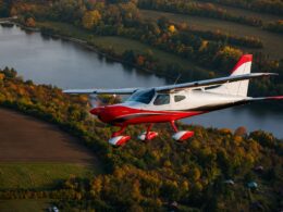 Red and white single engine plane in flight over autumn colored fields and a winding lake at sunset