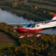 Red and white single engine plane in flight over autumn colored fields and a winding lake at sunset