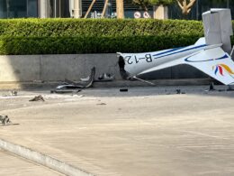 Small white airplane wrecked on a road with blue stripes tail damaged and scattered debris nearby